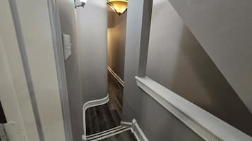Un pasillo estrecho con piso de madera, zócalo blanco y un aplique en la pared.