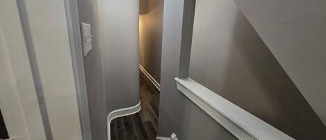 Un pasillo estrecho con piso de madera, zócalo blanco y un aplique en la pared.