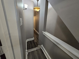 Un pasillo estrecho con piso de madera, zócalo blanco y un aplique en la pared.