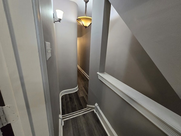 A narrow hallway with a wooden floor, a white baseboard, and a wall-mounted light fixture. - Charming 6 Bedroom & 4 Bath Home Near Philadelphia (Camden)