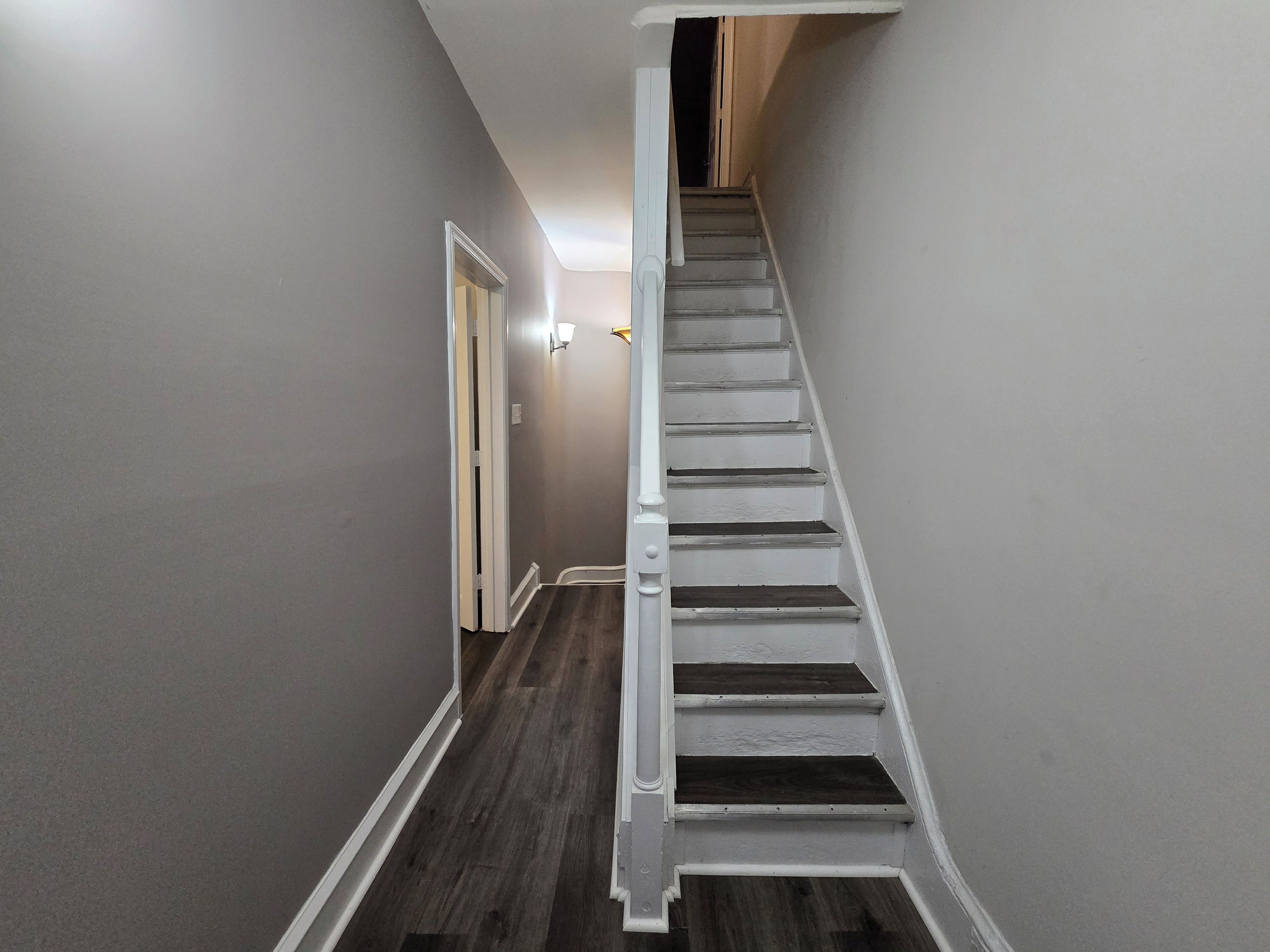 A narrow hallway with a wooden floor leading to a staircase with white railings.