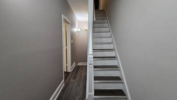 A narrow hallway with a wooden floor leading to a staircase with white railings.