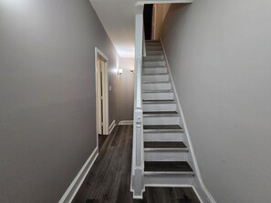 A narrow hallway with a wooden floor leading to a staircase with white railings. - Charming 6 Bedroom & 4 Bath Home Near Philadelphia (Camden)