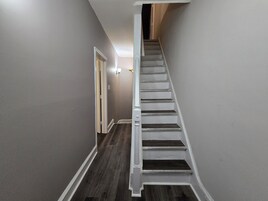 Un estrecho pasillo con piso de madera que lleva a una escalera con barandillas blancas.