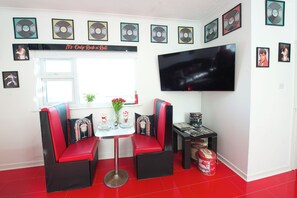 Ein Zimmer mit Fernseher, zwei roten und schwarzen Stühlen, einem kleinen Tisch mit einer Vase und gerahmten CDs an der Wand.