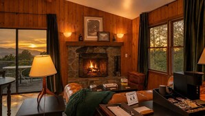 A cozy room with a fireplace, wooden walls, a view of the sunset, and a table with a lamp. - Coca-Cola Cabin @Fire Mountain Estate & Club. Endless views, Fire Pit, Jacuzzi! (Scaly Mountain)