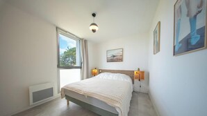 Et soveværelse med seng, et vindue, en vægmonteret lampe og et billede.