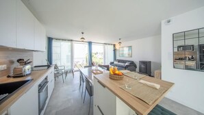 Et moderne køkken med en central ø, et spiseområde med bord og stole, samt en opholdsstue med sofa og fjernsyn.