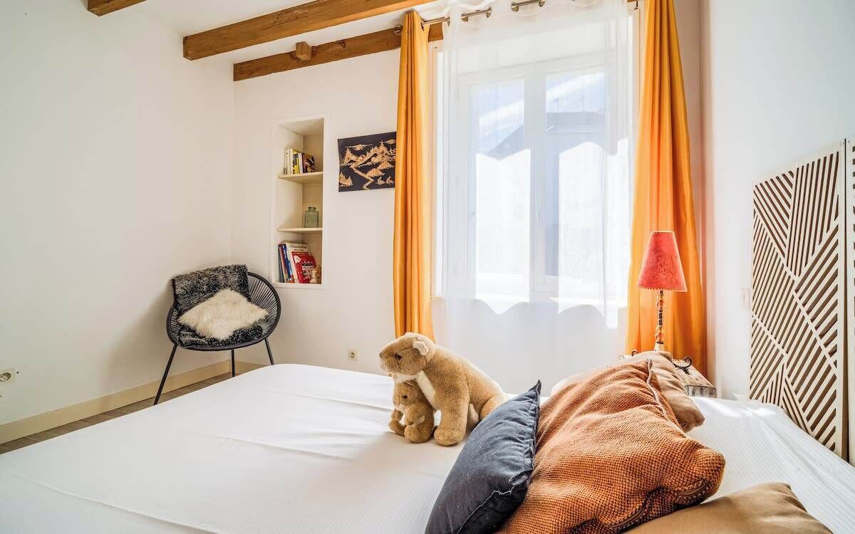 Une chambre à coucher avec un lit, une chaise, un ours en peluche, une lampe et une fenêtre avec des rideaux.