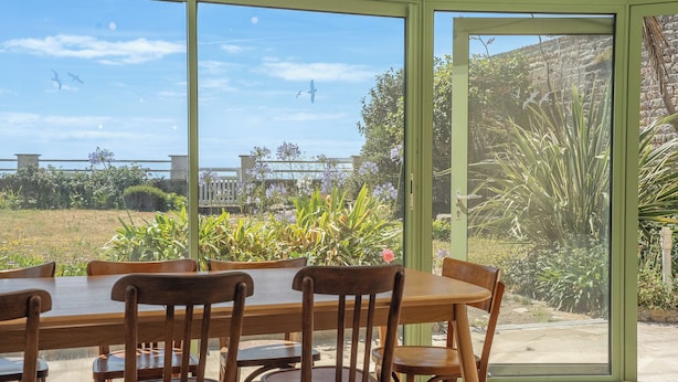 A dining area with a table and chairs, a large window with a view of a garden and a building. - Le Grand Large : Maison de Maître face mer (Penmarch)