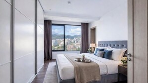 Una habitación de hotel moderna con una cama grande, una mesita de noche, un ventanal y una vista urbana a través del mismo.