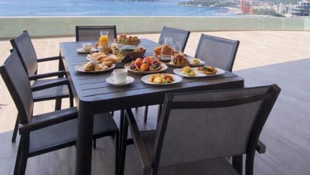 Una mesa de comedor con sillas, platos de comida y vistas al mar y a las colinas.