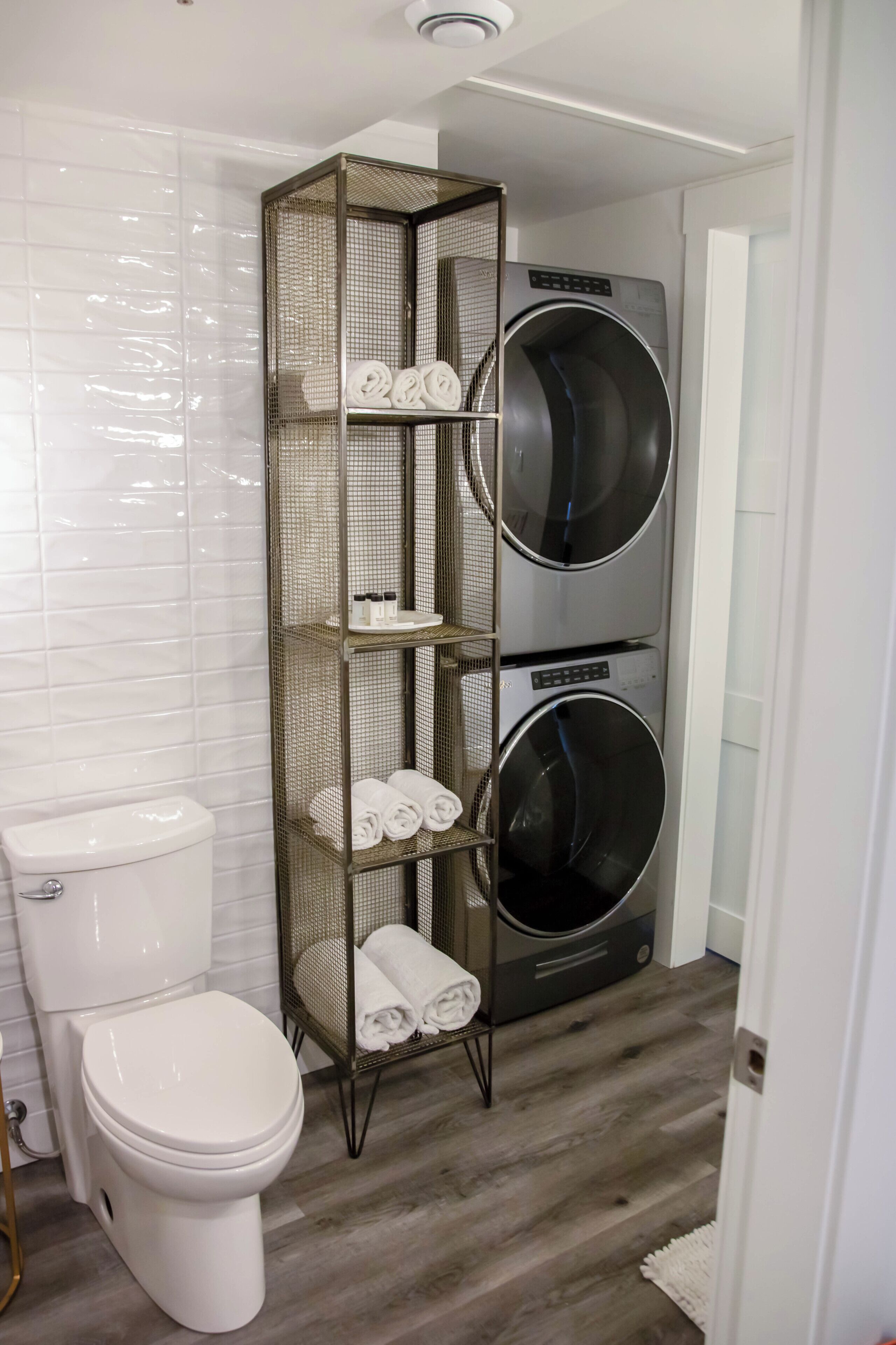 A bathroom with a white toilet, a metal laundry stand with folded towels, and a front-loading washing machine.