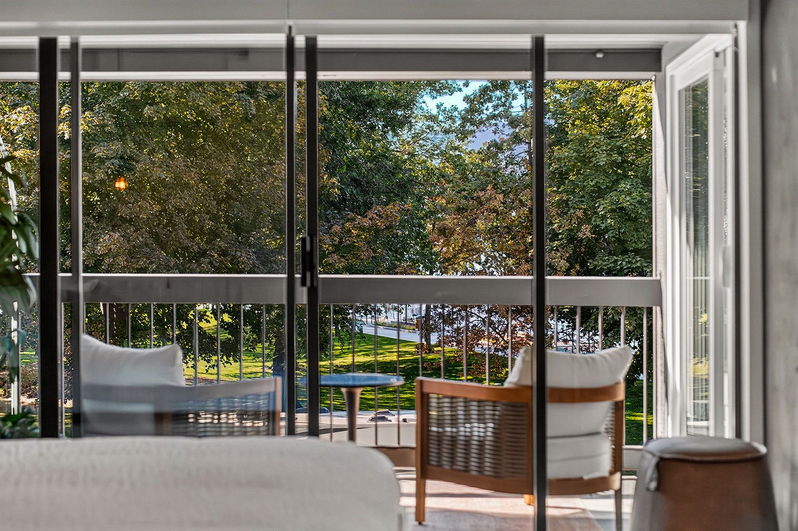 A balcony with a view of a garden and trees, a bed with white bedding, and two wicker chairs.
