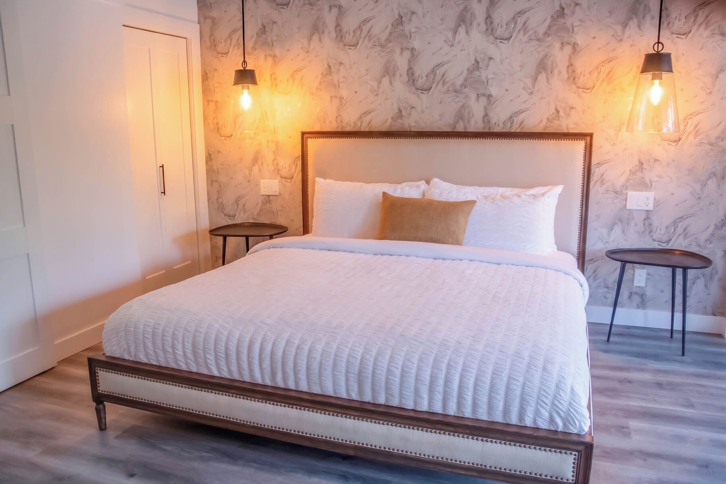 A neatly made bed with a wooden frame and white bedding, two bedside tables, and a marble-patterned wall with two hanging light fixtures.