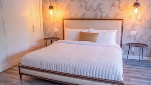 A neatly made bed with a wooden frame and white bedding, two bedside tables, and a marble-patterned wall with two hanging light fixtures. - LUXURY DOWNTOWN WATERFRONT RETREAT with Park, Lake and Marina Views (Kelowna)