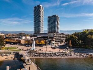 Un paisaje urbano con dos edificios altos destacados, una zona costera con un muelle y un parque público con una escultura.