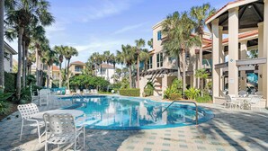 Una piscina con lettini e una zona salotto coperta, circondata da palme e edifici.