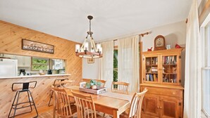 Een houten interieur met een eettafel, stoelen, een kroonluchter, een keukenhoek en een boekenkast.