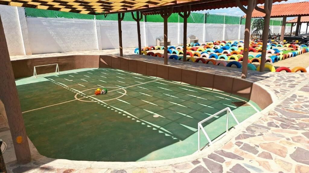 Una cancha deportiva interior con superficie verde, rodeada por un muro de piedra y una pérgola de madera.