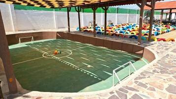 Una cancha deportiva interior con superficie verde, rodeada por un muro de piedra y una pérgola de madera.