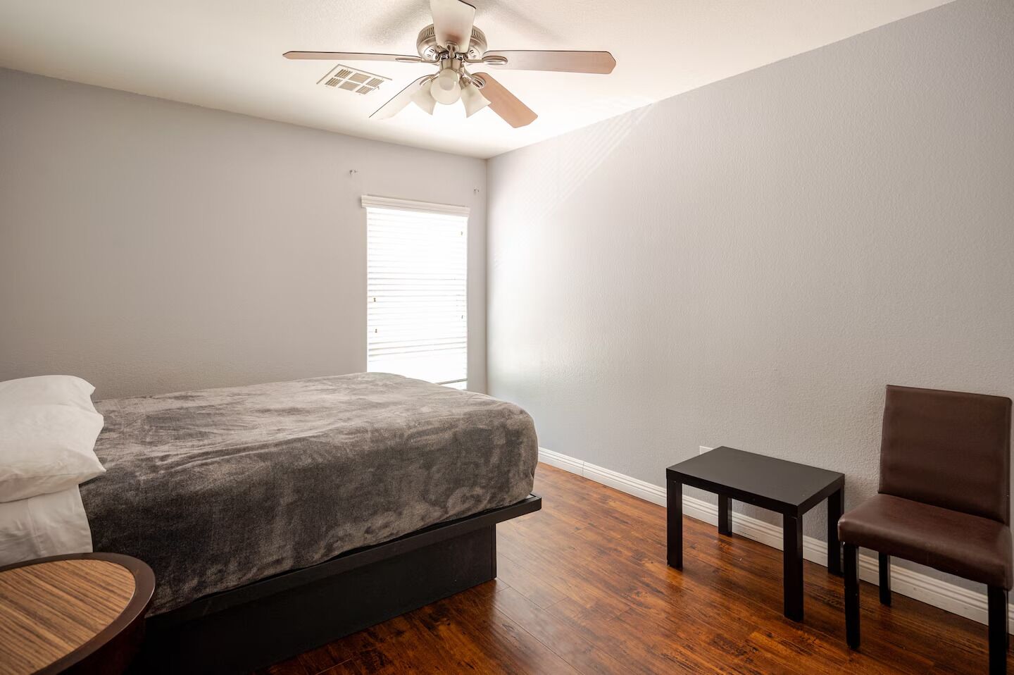 Une chambre à coucher avec un lit, une chaise, une petite table, un ventilateur de plafond et une fenêtre.