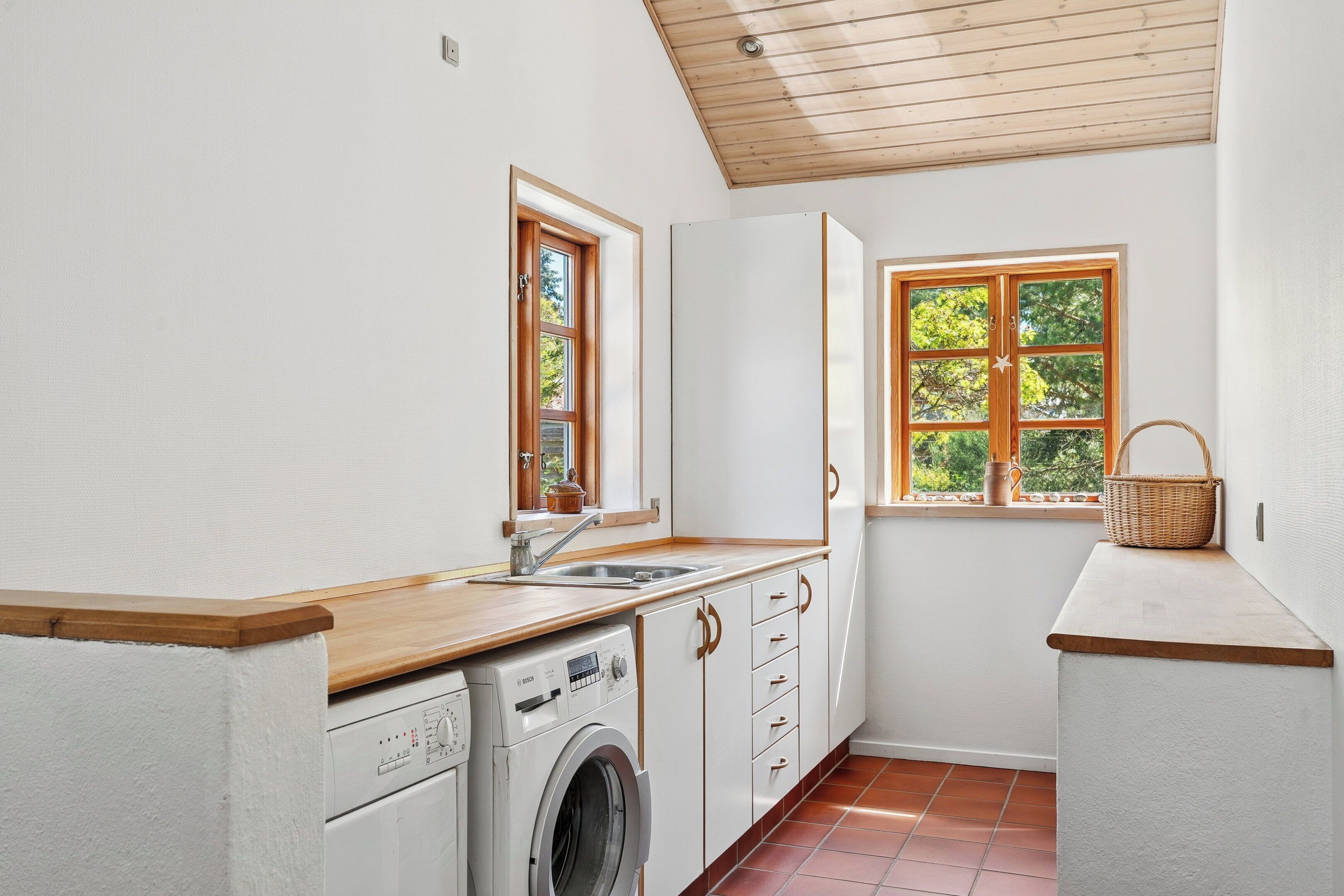 A laundry room with a washing machine, a sink, wooden countertops, and a window with a view of trees.