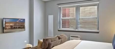 A bedroom with a bed, a chair, a small table, a window with blinds, and a wall-mounted picture.