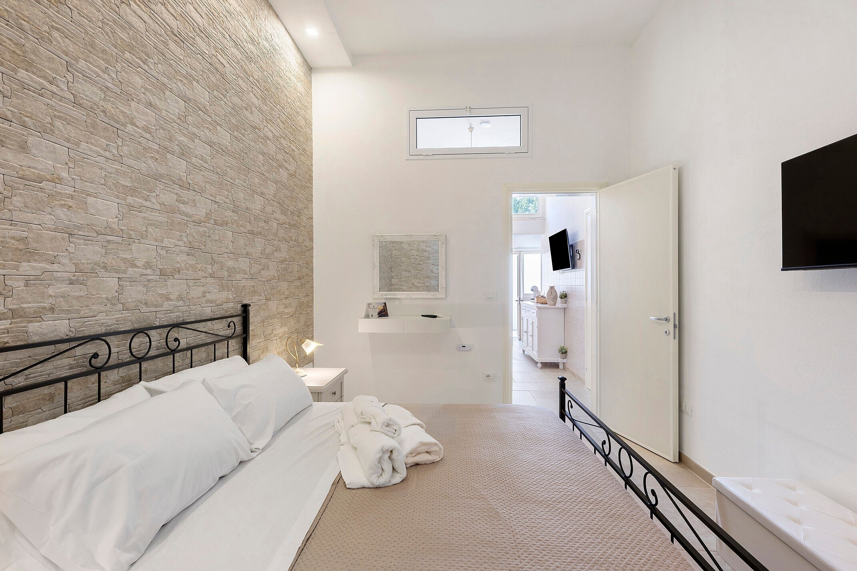 Un dormitorio con pared de piedra, una cama, una mesita de noche, un televisor y un baño visible a través de una puerta abierta.