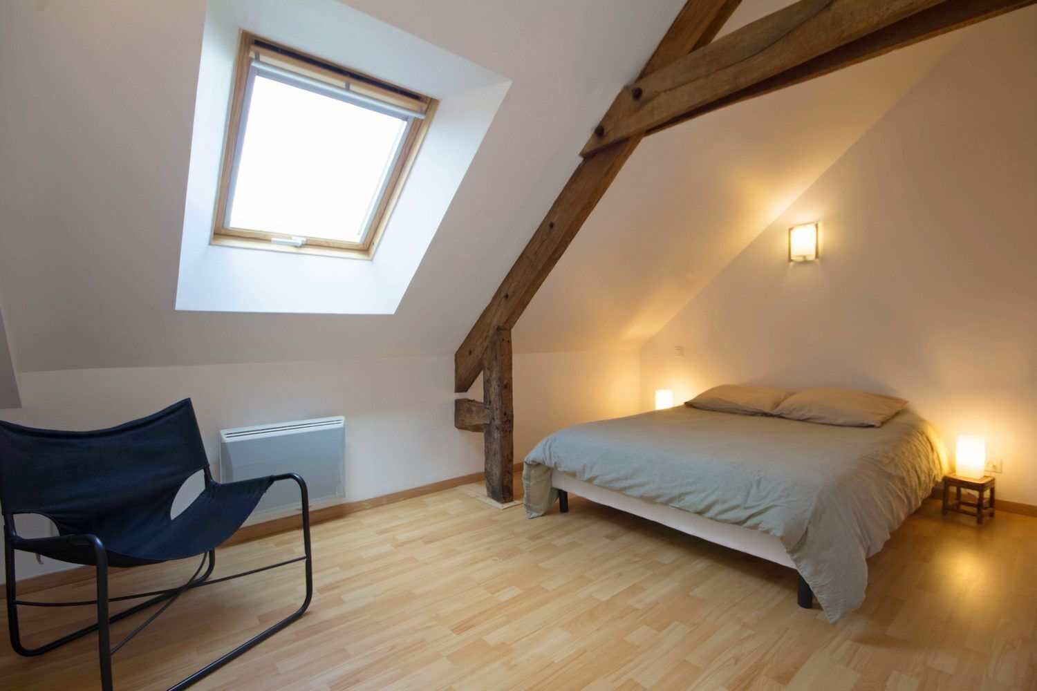 A bedroom with a sloped ceiling, a skylight, a bed, a chair, and a bedside table with a lamp.