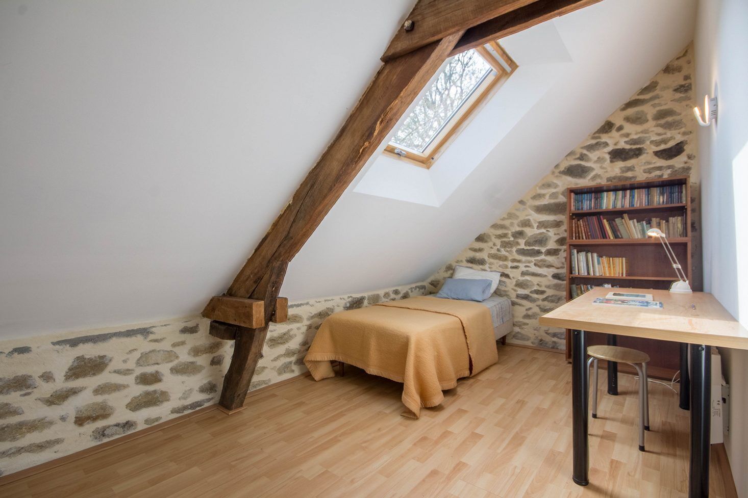 A cozy attic bedroom with a sloped ceiling, a wooden beam, a small bed, a desk with a chair, and a bookshelf.