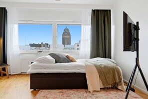 Ein Schlafzimmer mit einem Bett, einem Fenster mit Blick auf die Stadt, einem Fernseher auf einem Schrank und einem Holzboden.