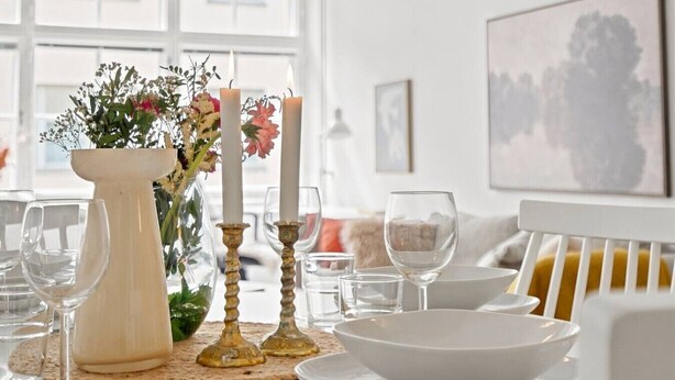 Ett matbord dukat med vita tallrikar, skålar och glas, en vas med blommor och ljus.
