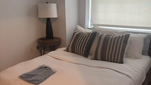 Ein Schlafzimmer mit einem Bett, Kissen, einer Lampe, einem Nachttisch und einem Fenster.