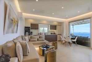 Ein modernes Wohnzimmer mit einer Couch, einem Couchtisch und Sesseln, eine Küche mit Schränken und Geräten, sowie ein großes Fenster mit Blick.