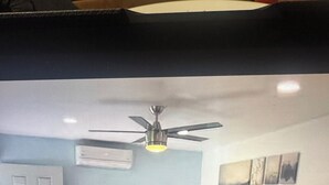 Ein Schlafzimmer mit Bett, Deckenventilator, Klimaanlage, Schreibtisch, Stuhl und Bildern an der Wand.