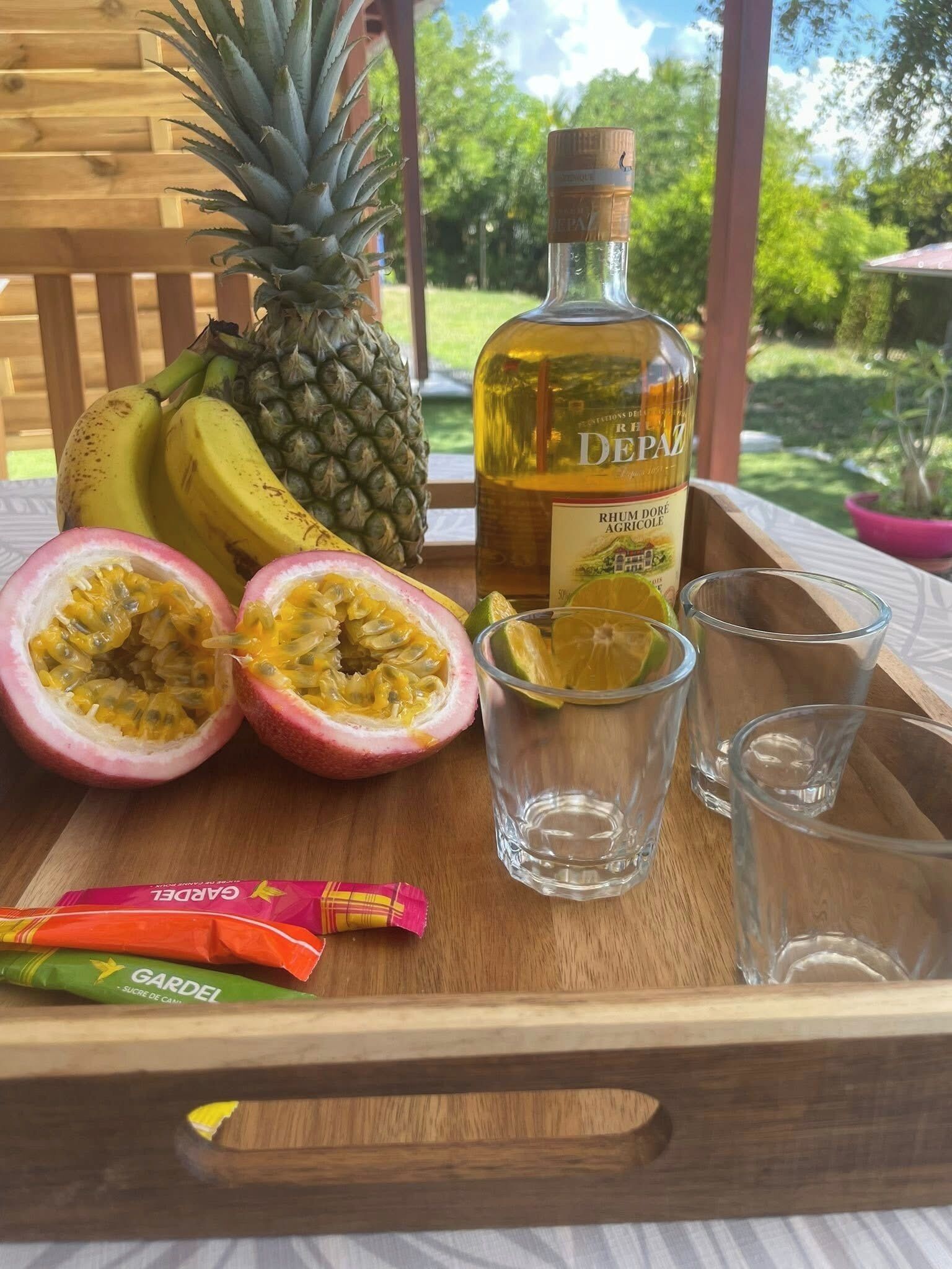 Una bandeja de madera con una botella de ron De Paz, una piña, plátanos, maracuyá y dos vasos para chupito.