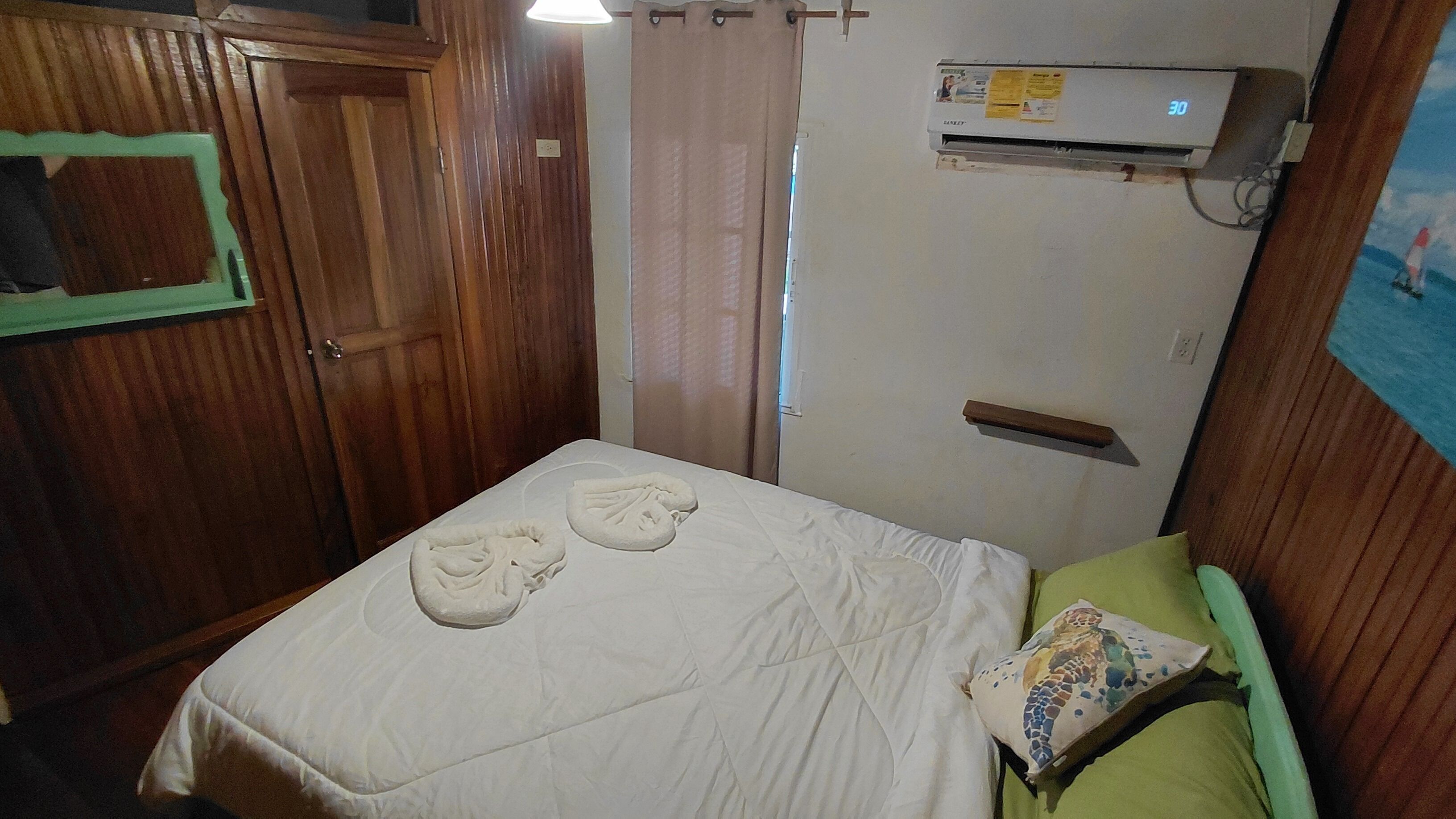 Ein kleiner, kompakter Raum mit einem Bett, einer Holztür, einer Wand-Klimaanlage und einem Vorhang.