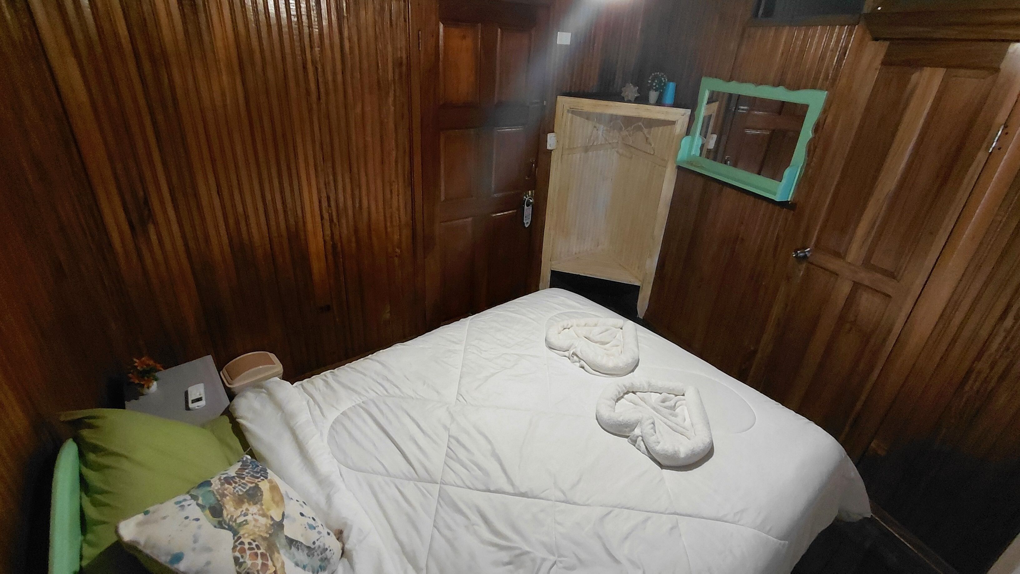 A room with wooden paneling, a bed with white bedding, a green pillow, a small bedside table with a phone, and a mirror on the wall.