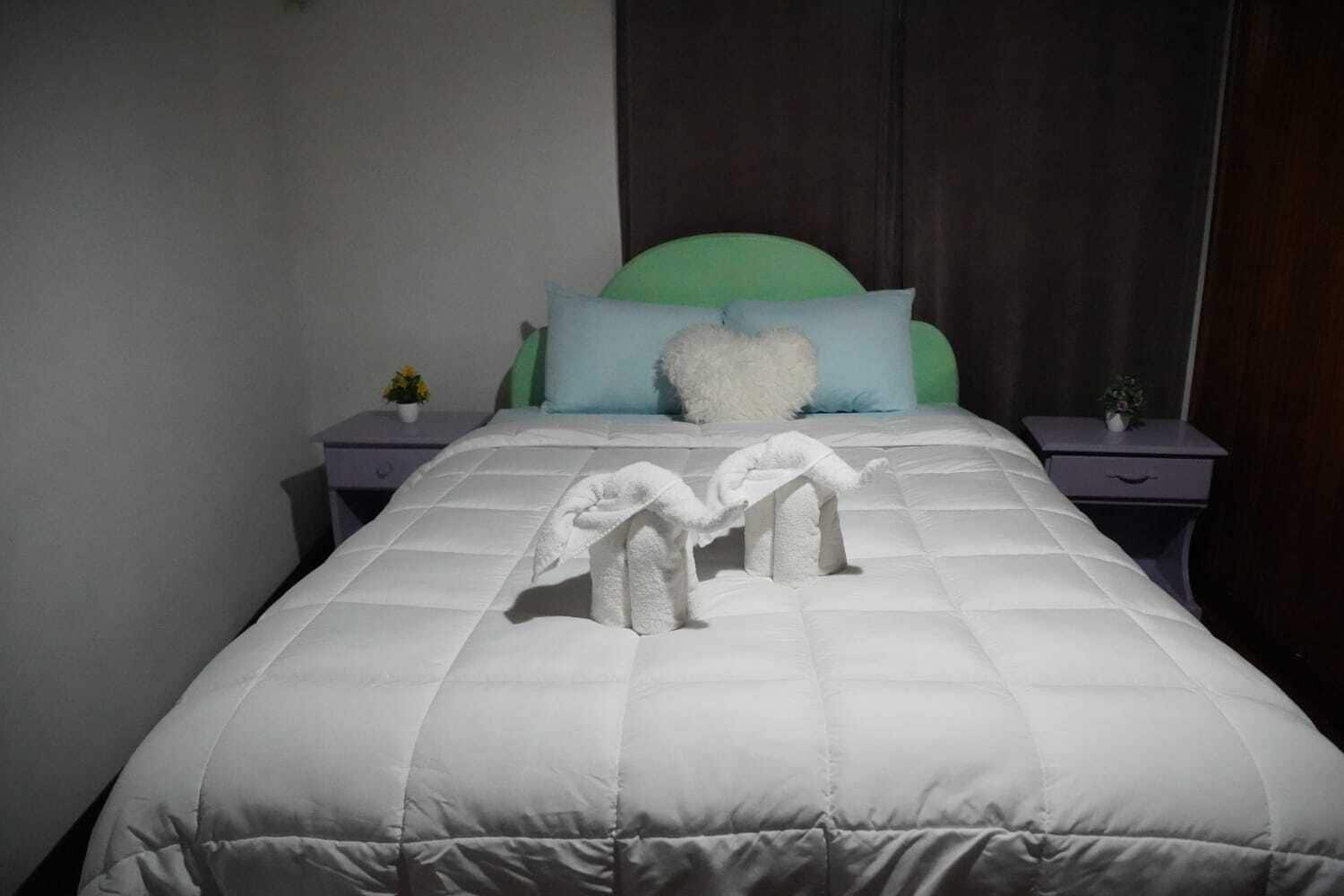 A neatly made bed with a white duvet, two towels arranged on the bed, and a green headboard.