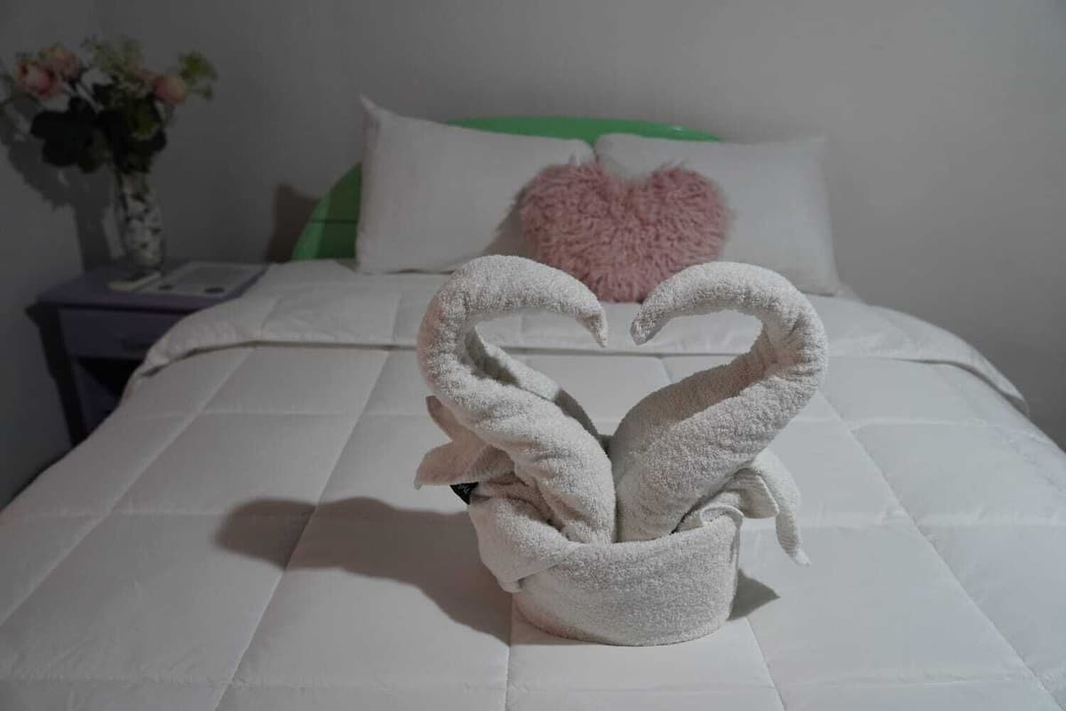 A bed with a heart-shaped towel arrangement, a white pillow with a green accent, and a vase with flowers on a nightstand.