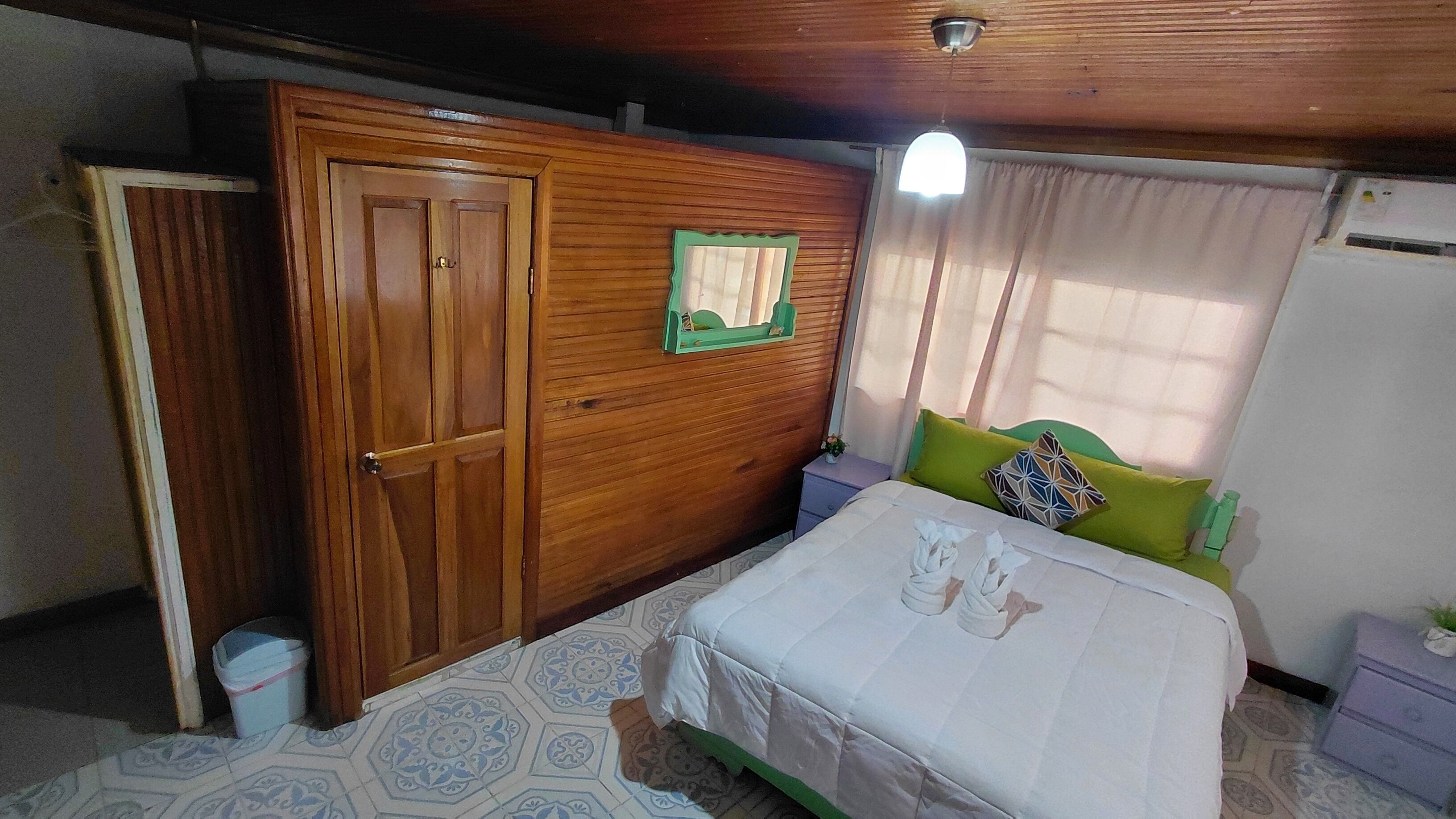 A bedroom with a wooden wardrobe, a bed with a white bedspread, a green pillow, and a nightstand with a lamp.