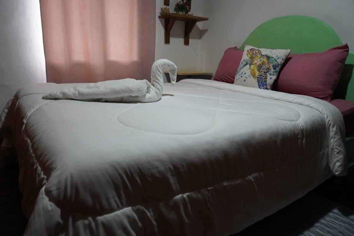 A neatly made bed with a white comforter and a towel, a green headboard, and a decorative pillow with a sea turtle design.