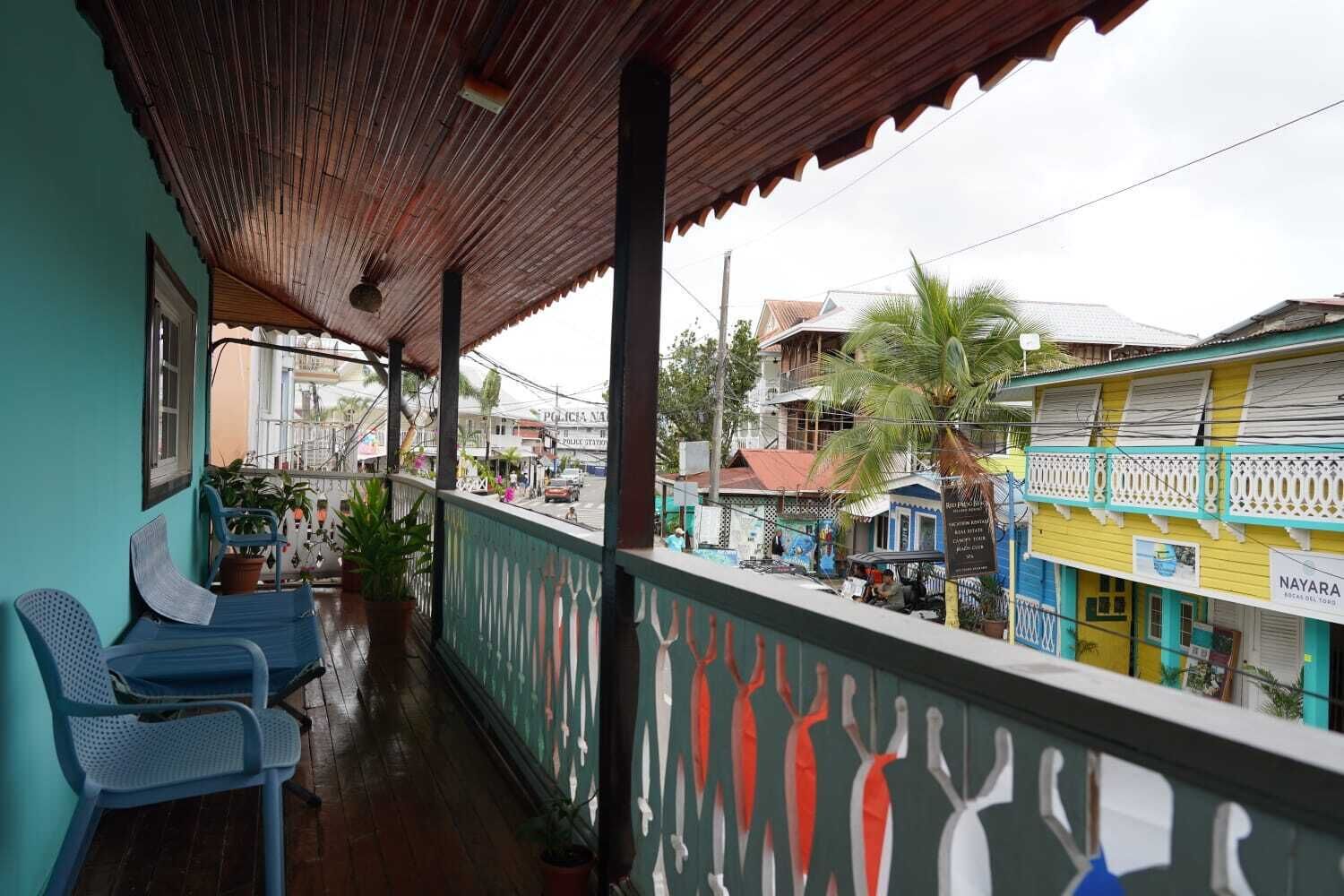 A covered balcony with chairs, a view of the street, and colorful buildings.
