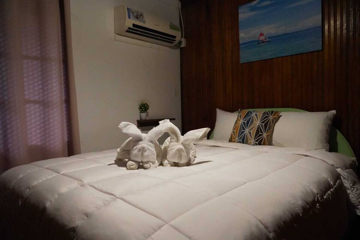 A hotel room with a bed, a painting of a sailboat, a wall-mounted air conditioner, and a small potted plant.
