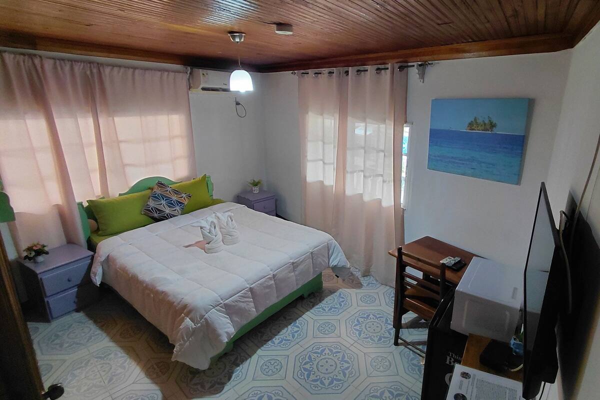 A bedroom with a bed, a desk, a chair, a TV, and a painting on the wall.