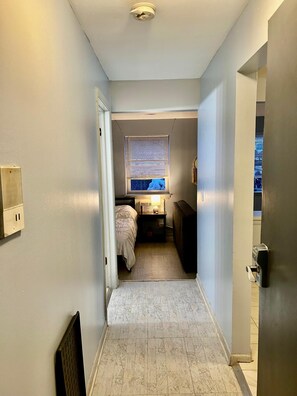 A narrow hallway leading to a bedroom with a bed, bedside table, and a window with blinds. - Awesome 1-bedroom studio with AC in vibrant Chicago (Chicago)