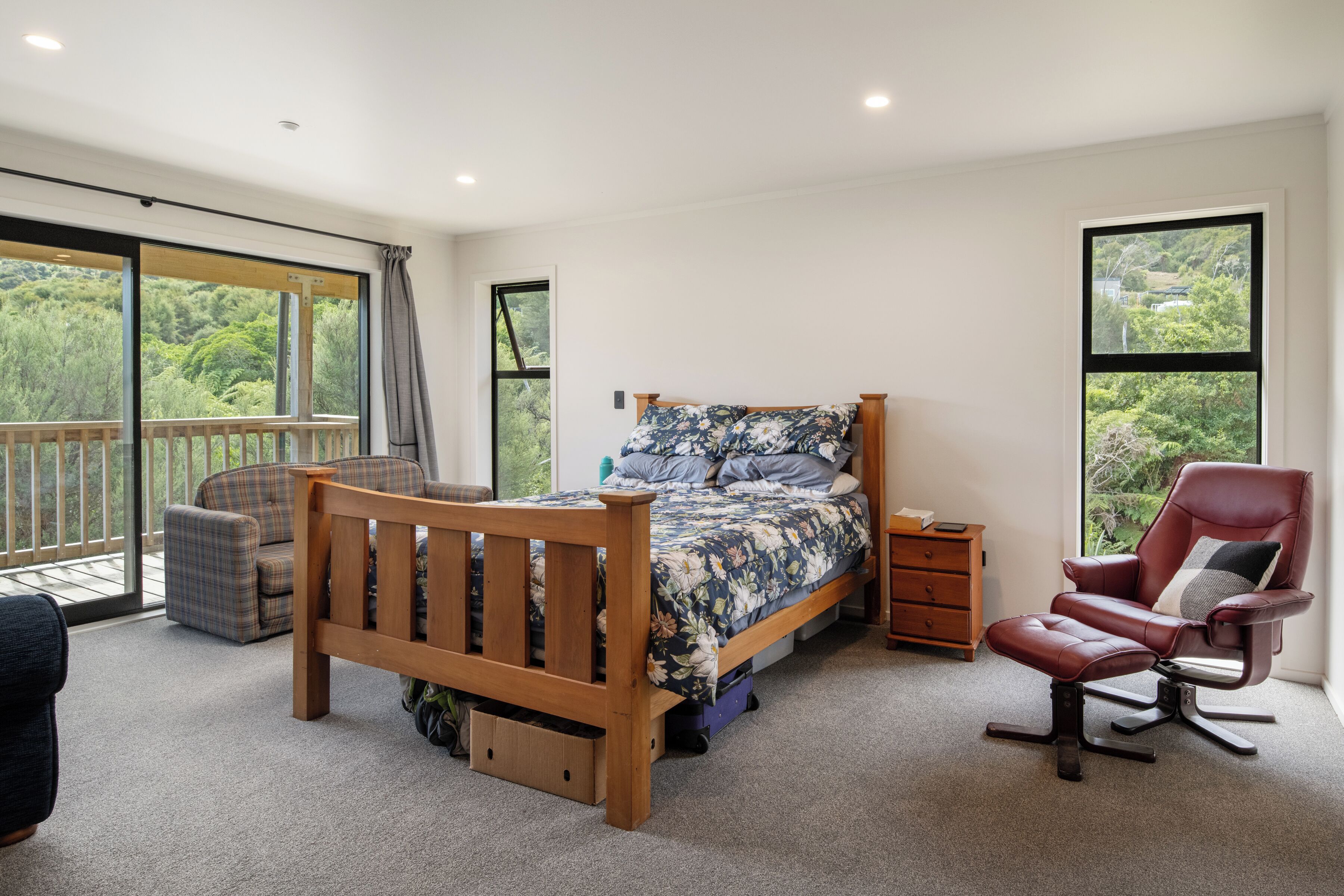 Um quarto com uma cama grande, cabeceira de madeira, uma cadeira, uma mesa pequena e vista para o exterior pela janela.