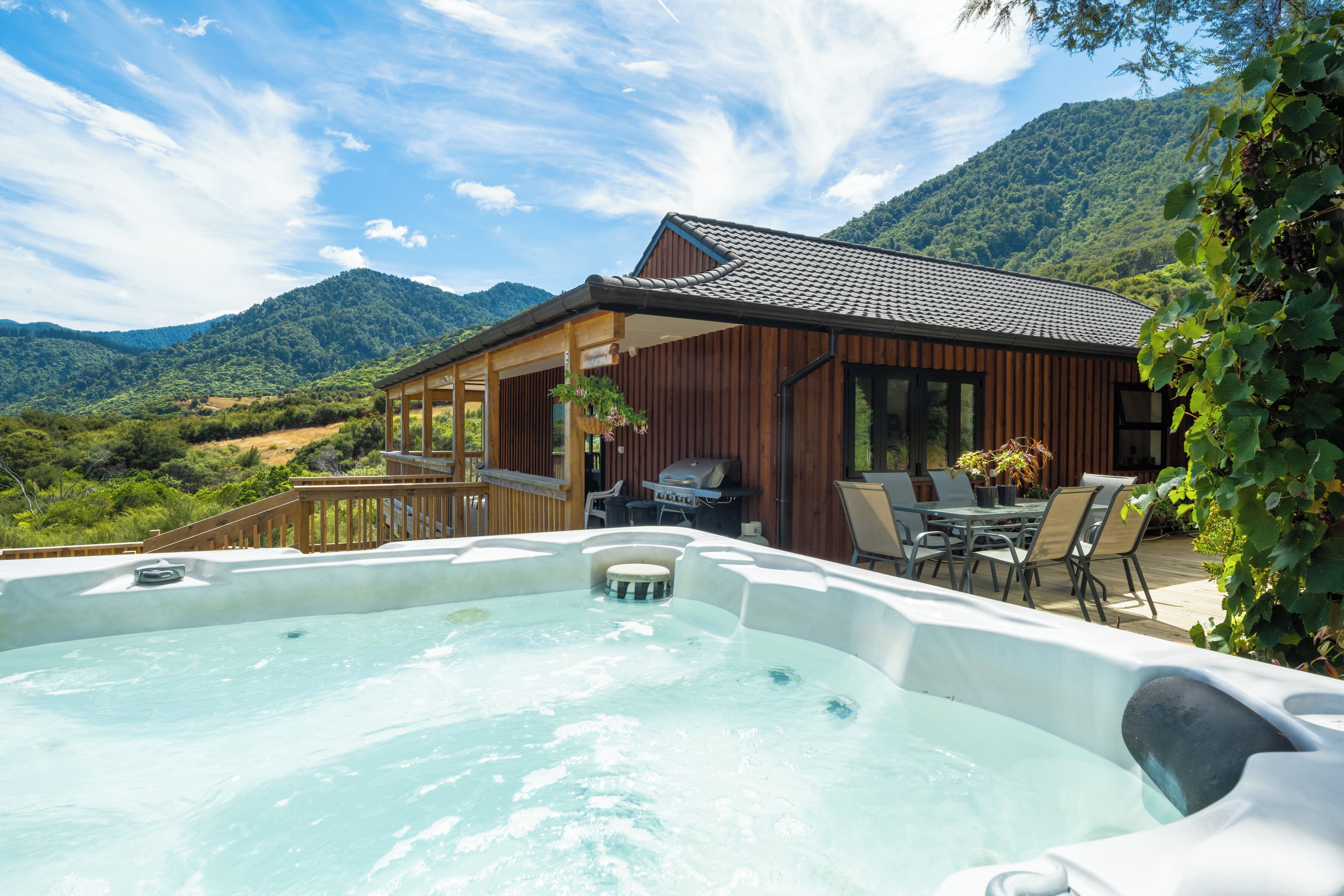 A hot tub with a wooden deck and outdoor furniture, surrounded by a mountainous landscape.