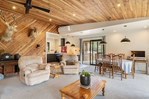 A spacious living area with a high ceiling, wooden beams, a fireplace, a dining set, and a kitchen visible through an open door.